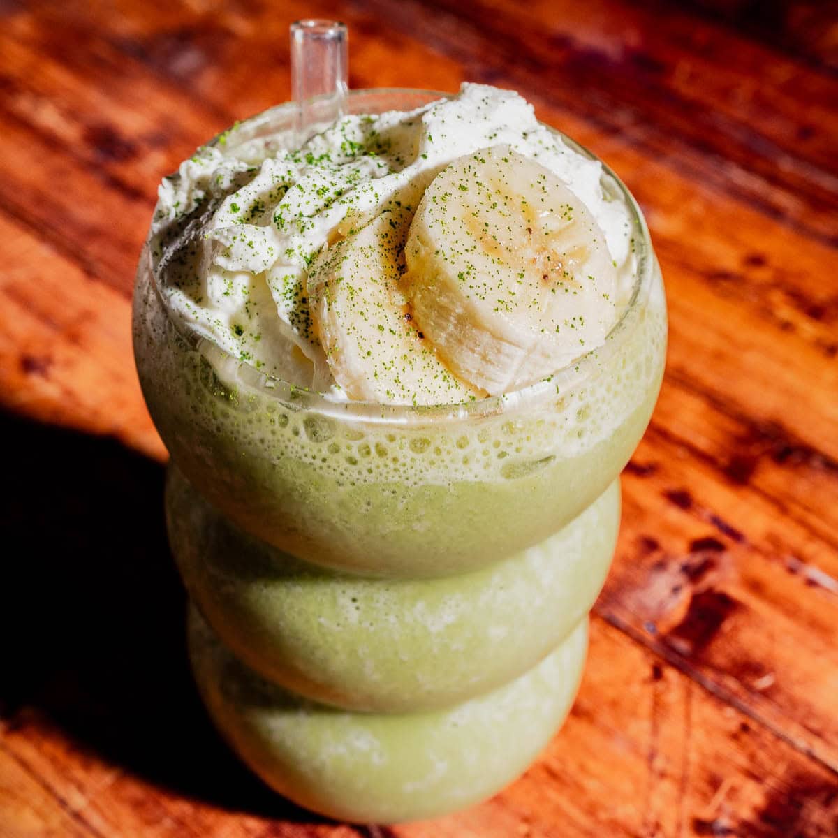 A matcha protein shake, topped with whipped cream, banana slices, and a sprinkle of green powder, served in a curvy glass on a wooden surface.