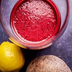 A glass of red beet pineapple juice seen from above, with a lemon and a whole beet placed next to the glass on a dark surface.