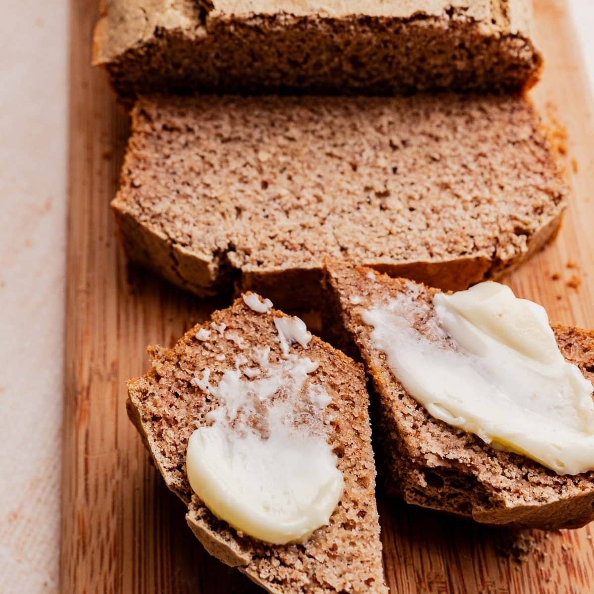 Sliced buckwheat bread on a wooden cutting board, with two pieces in the foreground spread with butter.