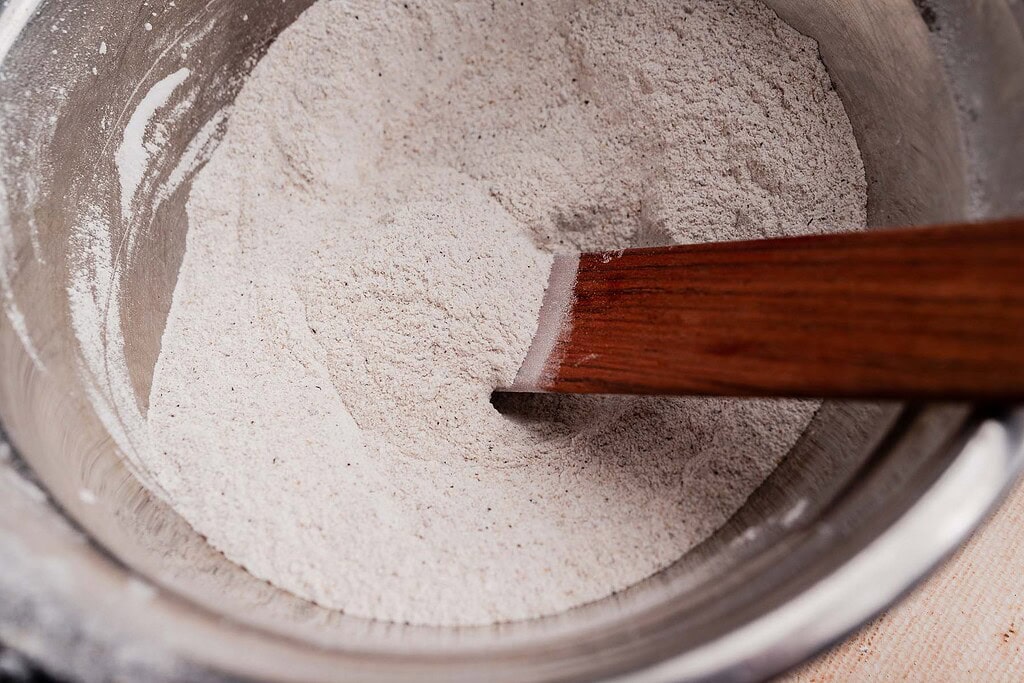 A metal mixing bowl containing dry flour for buckwheat bread is being stirred with a wooden spatula.