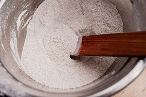 A metal mixing bowl containing dry flour for buckwheat bread is being stirred with a wooden spatula.