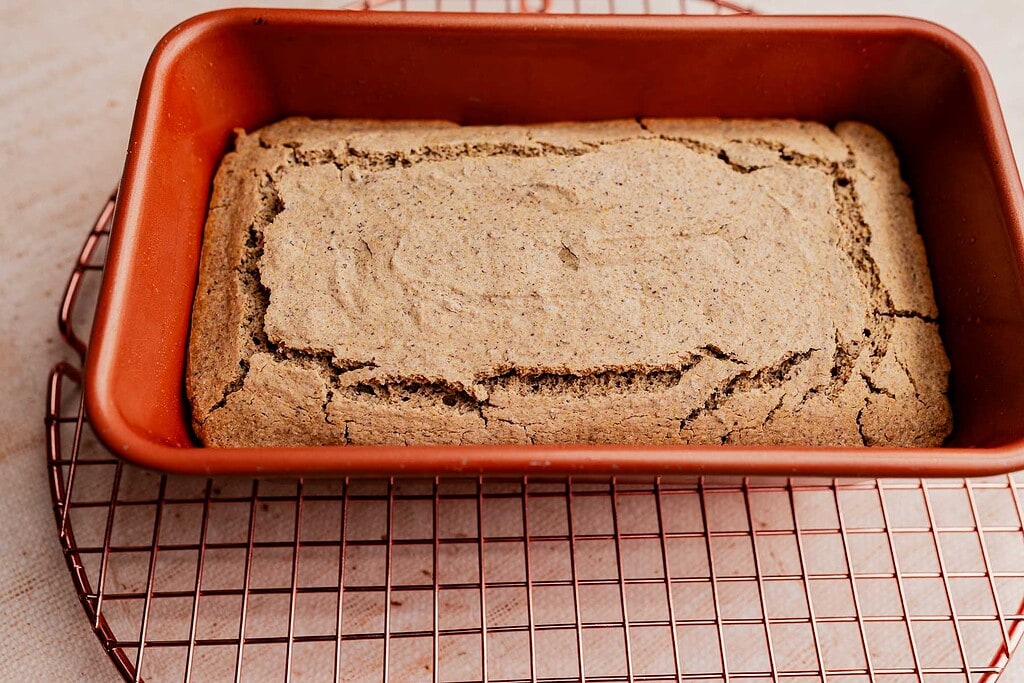 A loaf of buckwheat bread with a cracked top sits in a red silicone baking pan on a round wire cooling rack.