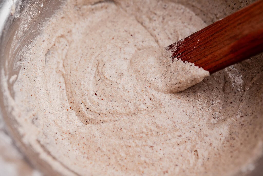 A wooden spatula stirs a thick, speckled buckwheat bread batter in a metal mixing bowl.