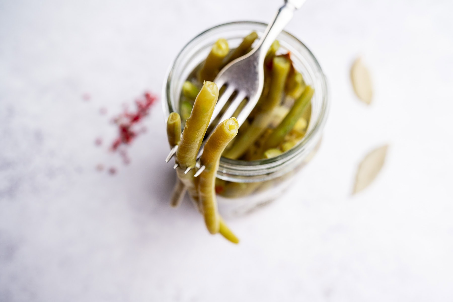 A fork lifting pickled green beans from a glass jar, with bay leaves and pink peppercorns scattered on a white surface, showcases the vibrant look of homemade pickled green beans.