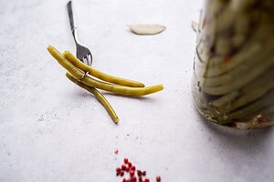 Three pickled green beans, pierced by a fork, sit on a white surface next to a glass jar of pickled green beans, with peppercorns and bay leaves scattered nearby.