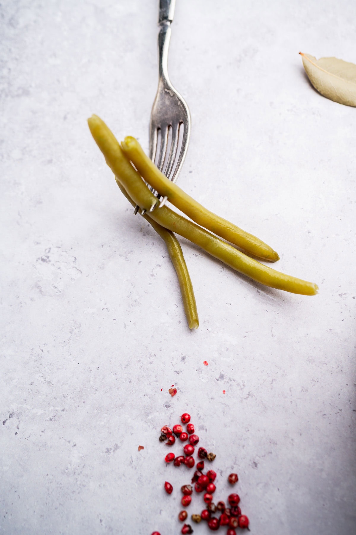 A fork holding three pickled green beans rests on a light surface with scattered red peppercorns and a bay leaf nearby.
