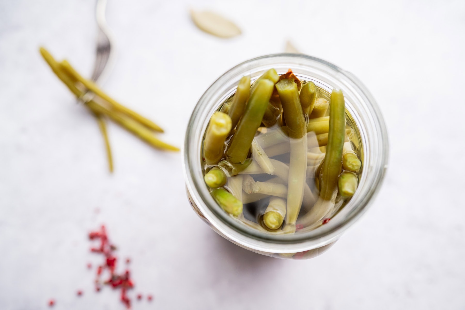A glass jar filled with pickled green beans in brine, viewed from above. Pickled green beans, a fork, bay leaf, and peppercorns are scattered on the white surface nearby.