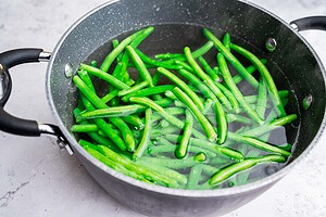 Pickled green beans are submerged in water inside a black pot on a light-colored surface.