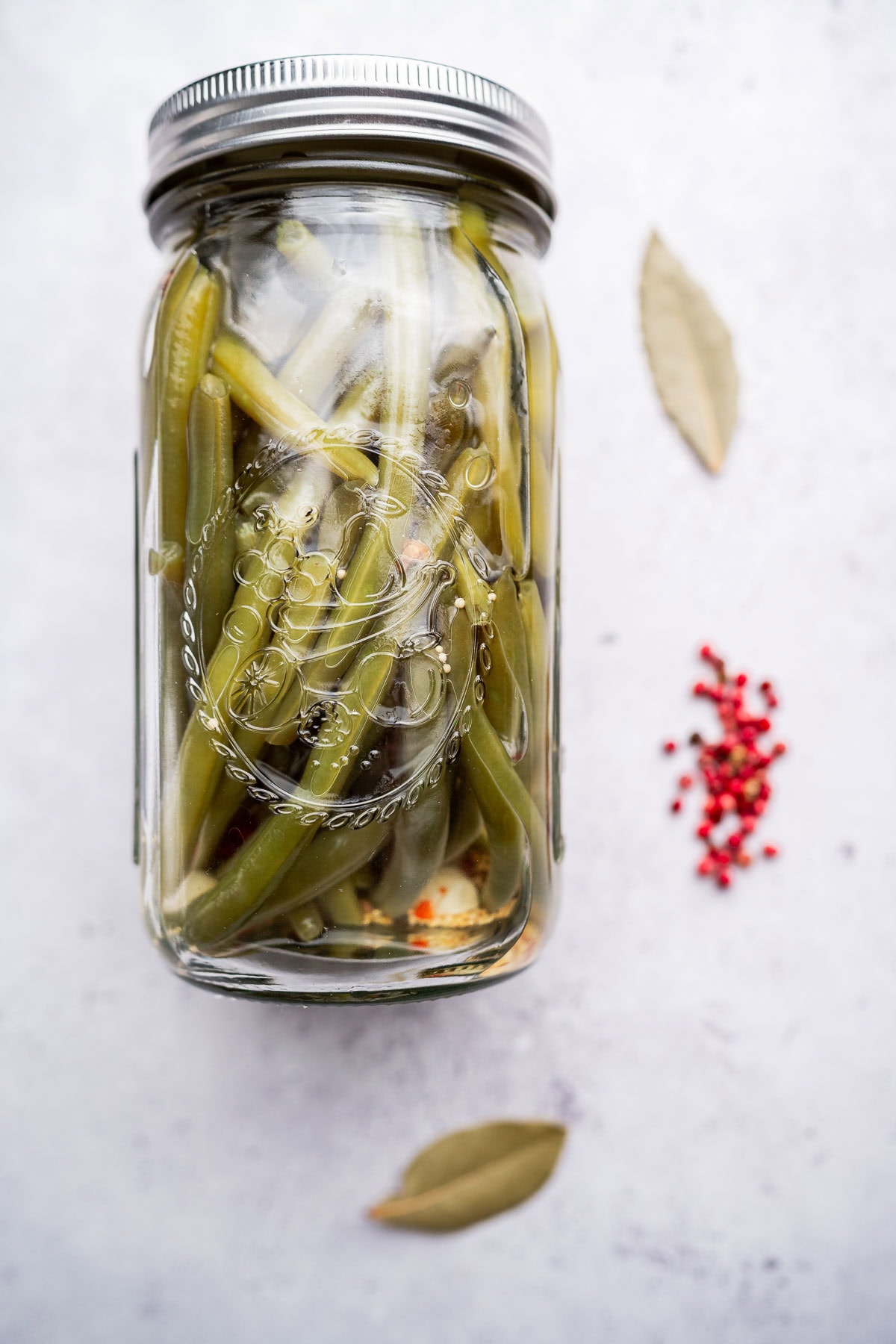 A glass jar of pickled green beans, sealed with a metal lid, rests on a light surface alongside bay leaves and scattered pink peppercorns.