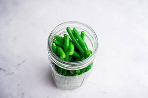 A glass jar filled with crisp pickled green beans sits on a light, textured surface.