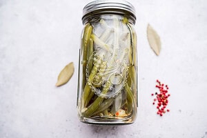 A glass jar of pickled green beans in brine sits on a white surface, with two bay leaves and a small pile of pink peppercorns arranged nearby.