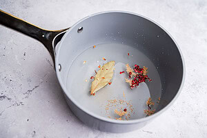 A gray saucepan with water, a bay leaf, mustard seeds, red peppercorns, and spice flakes—perfect for preparing pickled green beans—sits on a light-colored surface.