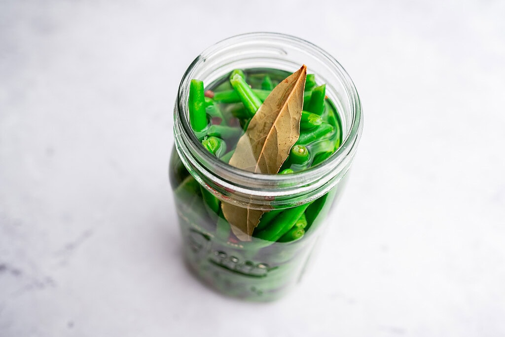 A glass jar filled with pickled green beans in liquid, topped with bay leaves, sits on a light surface.