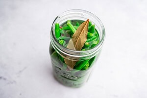 A glass jar filled with pickled green beans in liquid, topped with bay leaves, sits on a light surface.