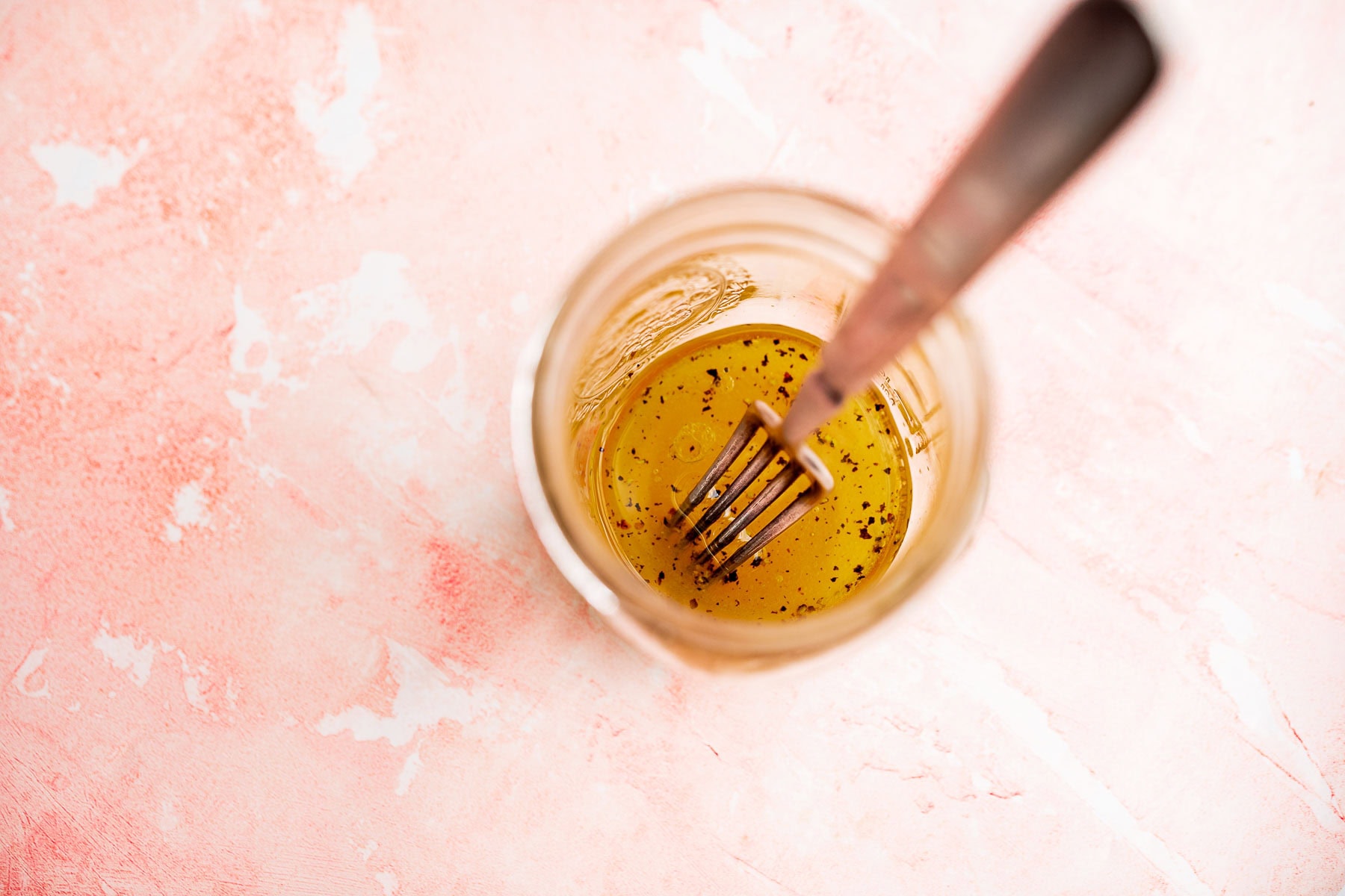 A glass jar with yellow salad dressing and black pepper specks, stirred with a metal fork, sits on a light pink textured surface&mdash;perfect for drizzling over a fresh radish salad.