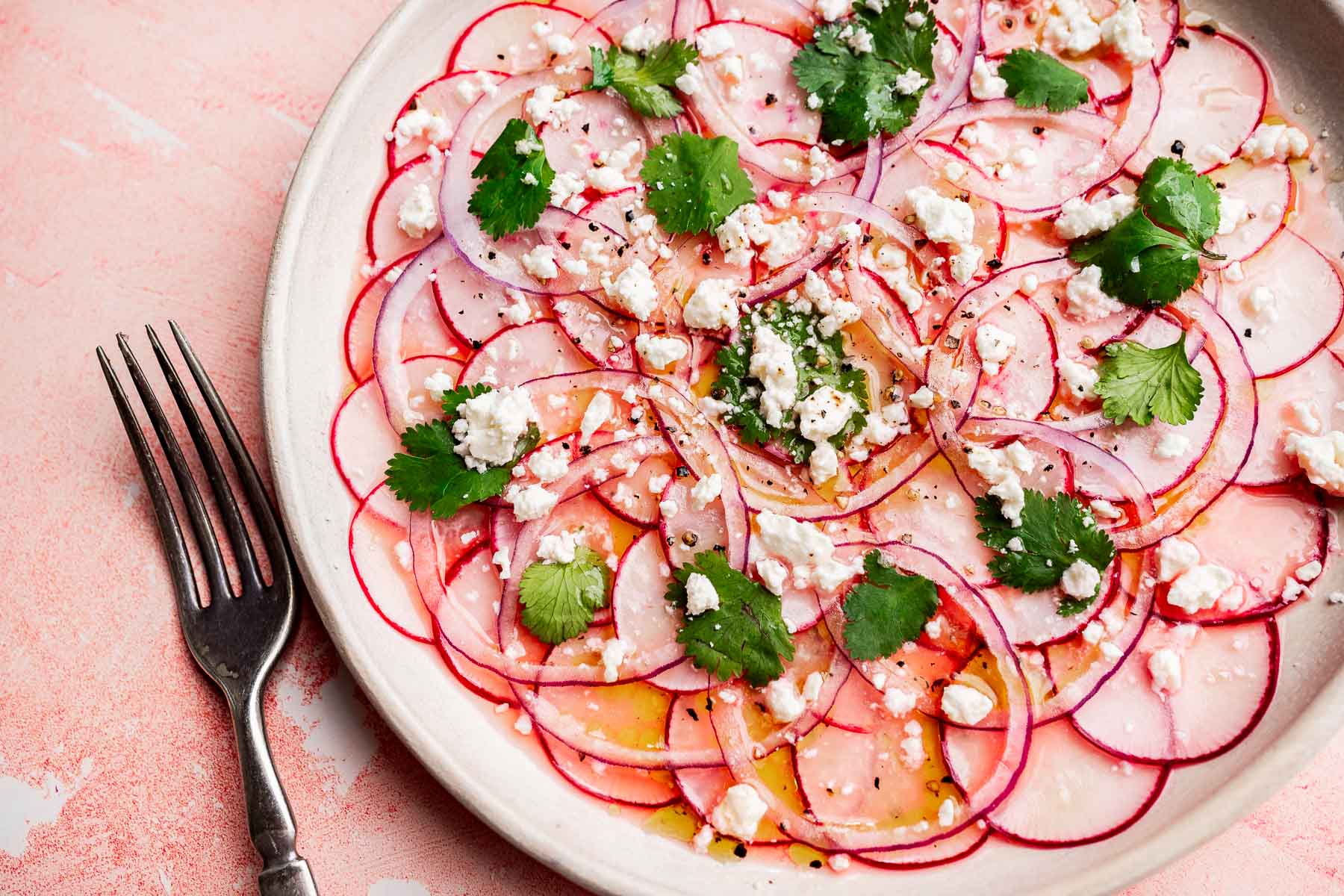 A vibrant radish salad featuring thinly sliced radishes topped with crumbled cheese, fresh cilantro leaves, and a drizzle of dressing, served with a fork beside the plate.