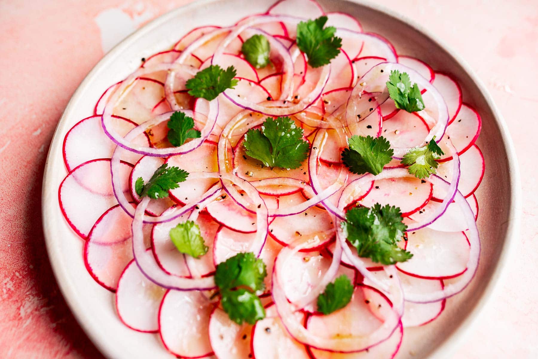 A vibrant radish salad featuring thinly sliced radishes and red onions, artfully arranged in a circular pattern, topped with fresh cilantro leaves and a light vinaigrette.