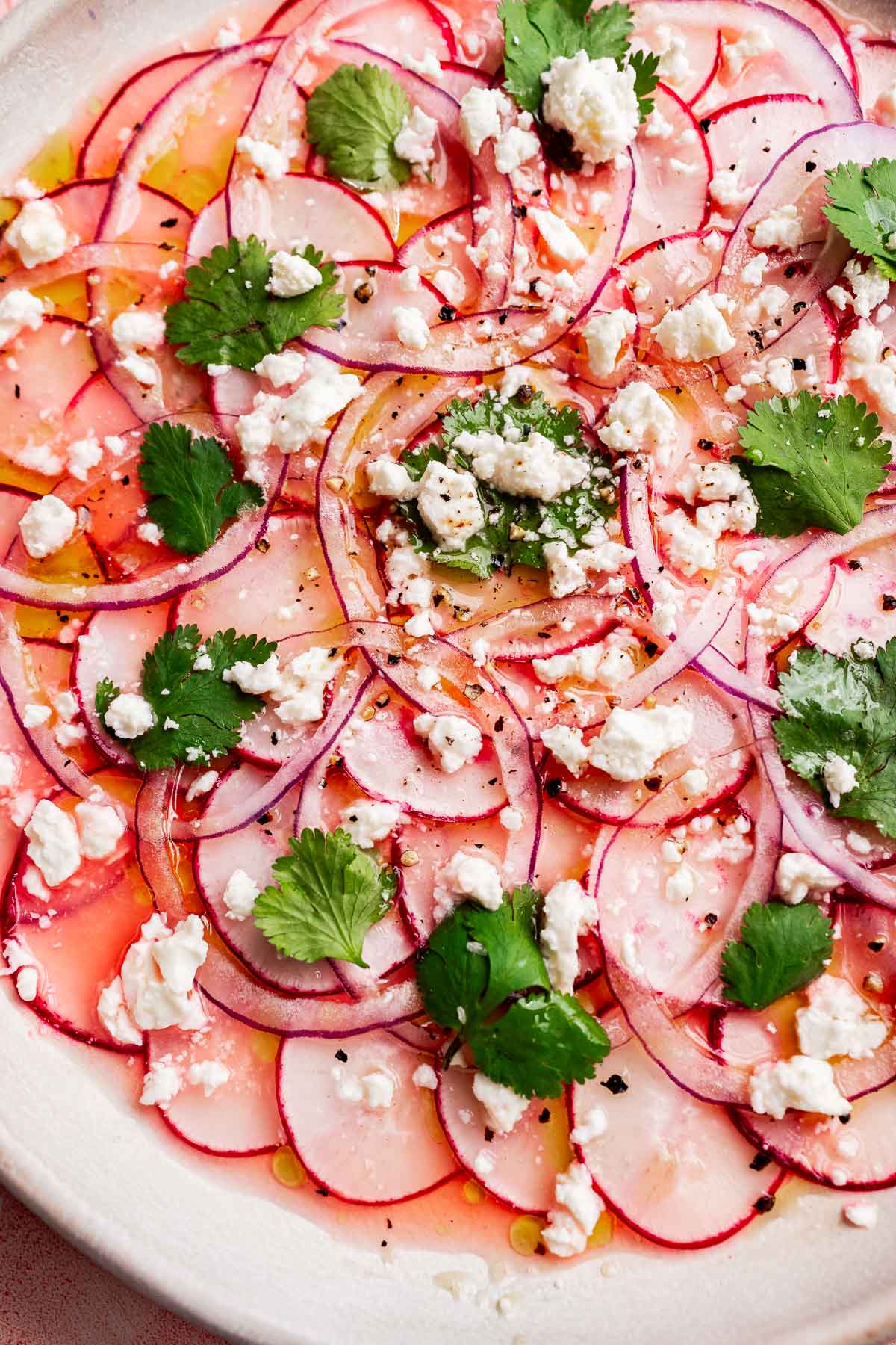 Thinly sliced radishes and red onions create a vibrant radish salad, topped with crumbled feta cheese, fresh cilantro leaves, black pepper, and a drizzle of olive oil on a white plate.