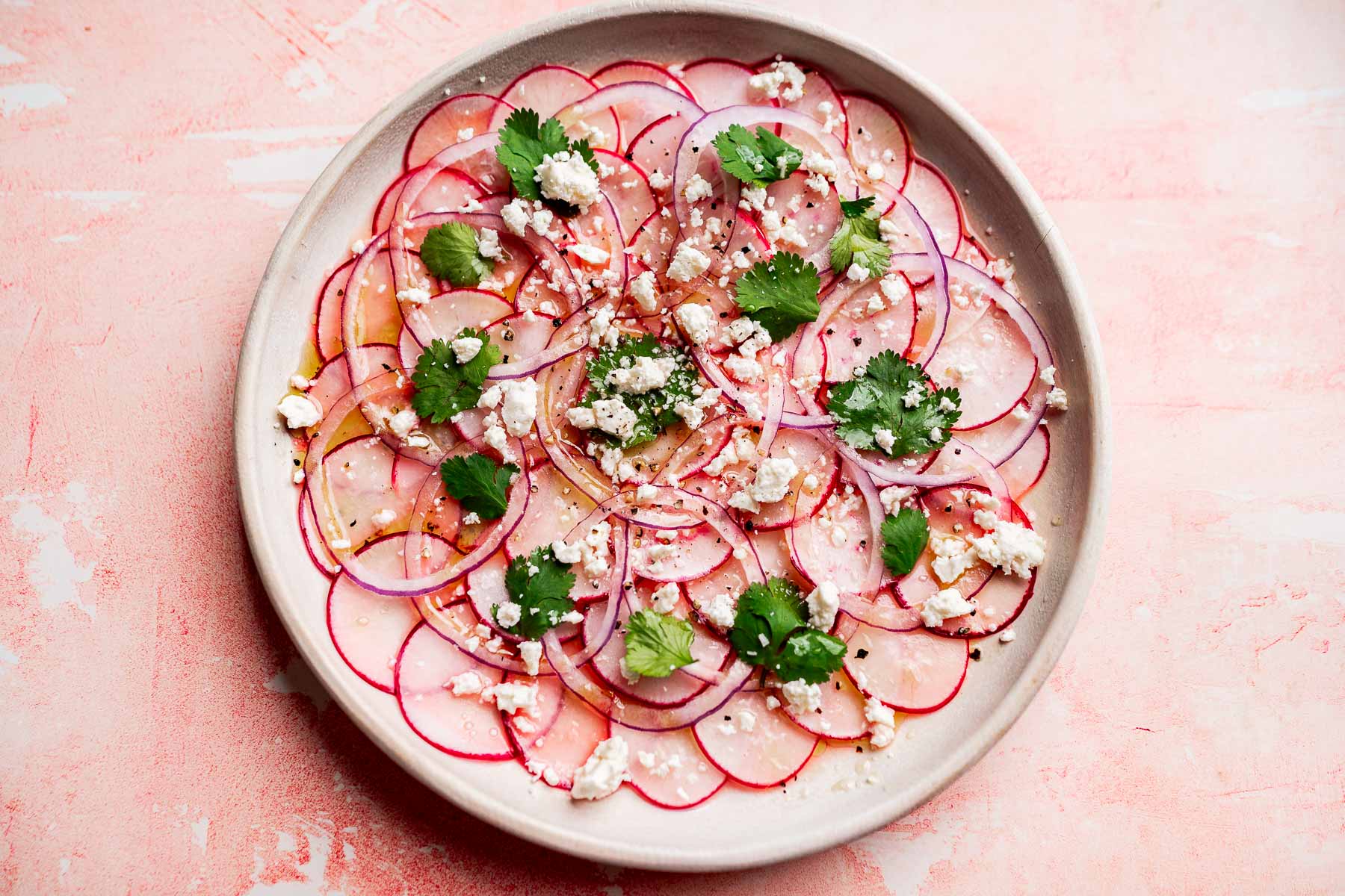 A vibrant radish salad featuring thinly sliced radishes arranged in circles, topped with crumbled feta cheese, red onion, cilantro leaves, and a drizzle of dressing on a light pink background.