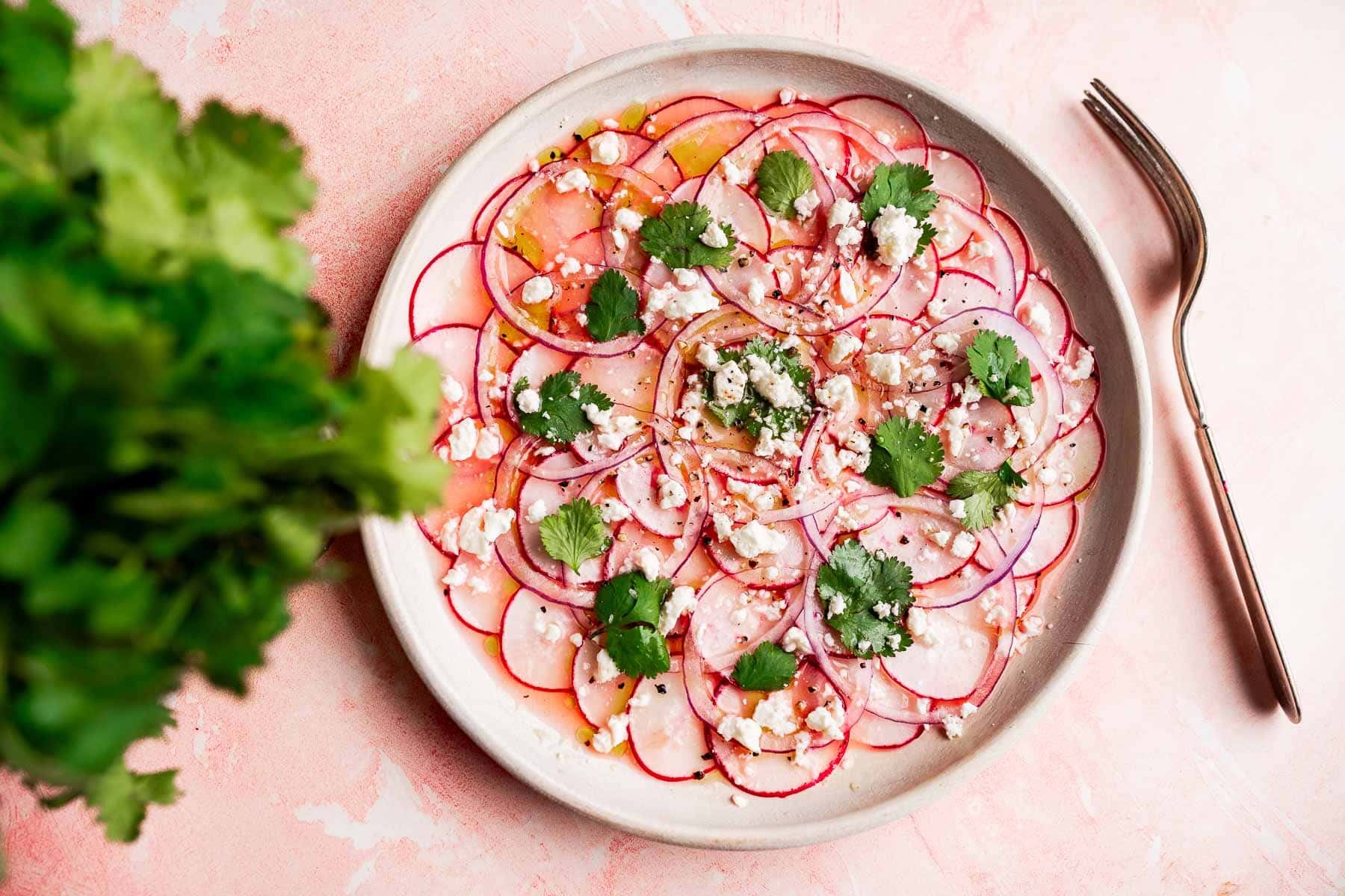 A vibrant radish salad featuring thinly sliced radishes topped with crumbled cheese, cilantro leaves, and seasoning, served on a pink surface with a fork and fresh cilantro nearby.
