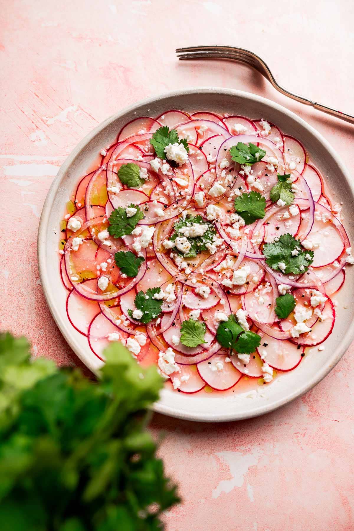 A vibrant radish salad featuring thinly sliced radishes topped with crumbled cheese, cilantro leaves, and a drizzle of dressing sits on a pink surface next to a fork.