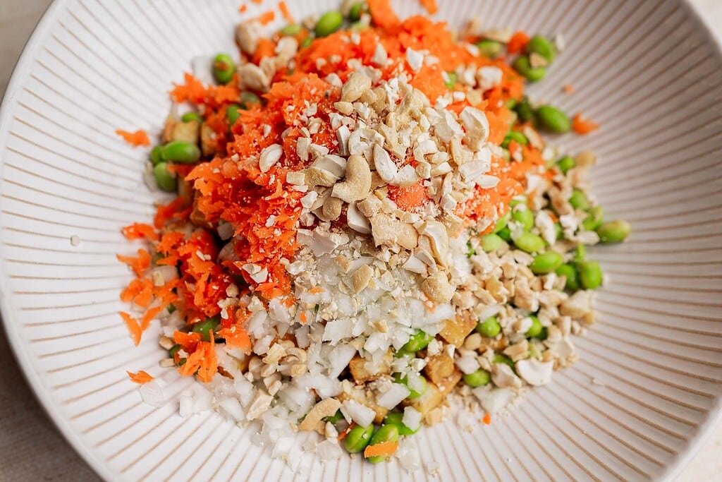 A close-up of a bowl featuring chopped carrots, onions, tofu, edamame, and crushed cashews arranged in separate piles—perfect for a vibrant tofu edamame salad.