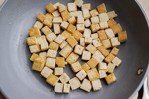 Cubed tofu pieces are being pan-fried in a non-stick skillet, lightly browned on some sides—perfect for adding to a refreshing tofu edamame salad.