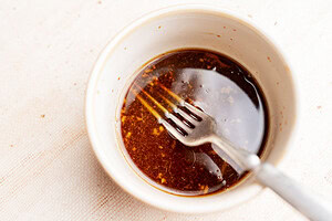A small white bowl containing a dark liquid sauce with visible spices, perfect for drizzling over tofu edamame salad, with a metal fork resting inside the bowl.