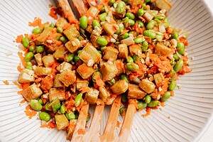 A close-up of a vibrant tofu edamame salad, featuring diced tofu, edamame, shredded carrots, and chopped nuts on a white plate with wooden utensils.