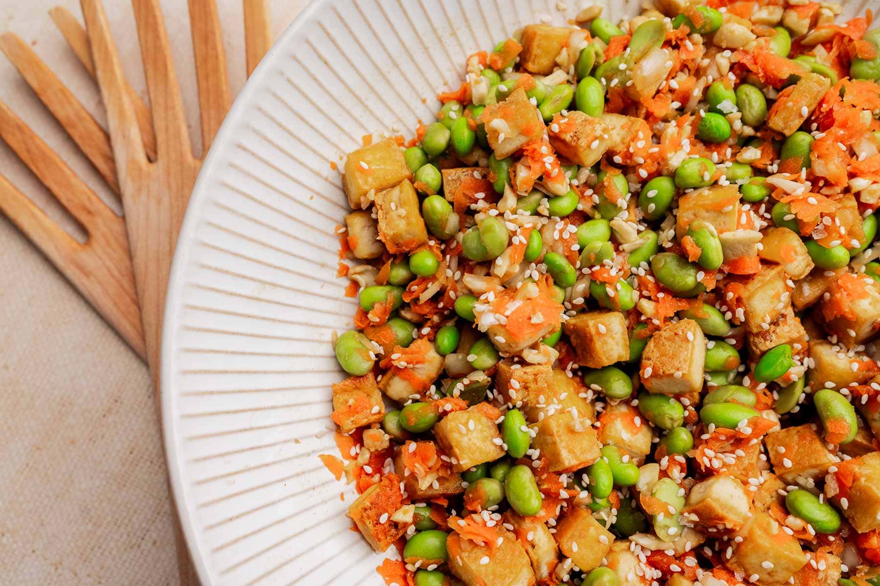 A white plate filled with a tofu edamame salad featuring cubed tofu, edamame, shredded carrots, and sesame seeds, with wooden forks beside the plate.