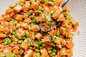 A close-up of a tofu edamame salad bowl with diced tofu, edamame, shredded carrots, and sesame seeds, as a spoon scoops up the vibrant mixture.