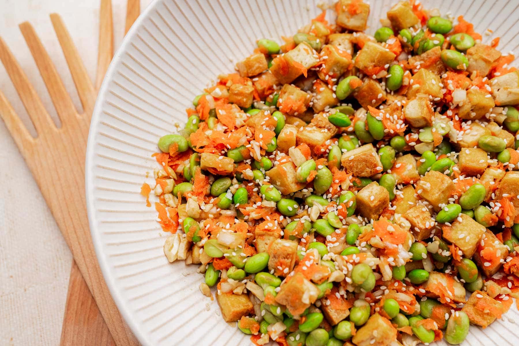 A vibrant tofu edamame salad featuring cubed tofu, edamame, shredded carrots, sesame seeds, and grains, served in a white dish with wooden utensils beside it.