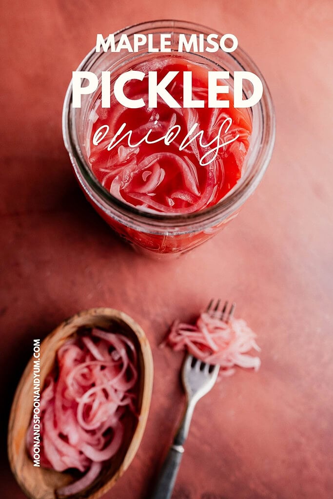 A jar of miso pickled onions with a hint of maple sits on a pink surface, while tangy onions rest in a small wooden bowl and on a fork beside the jar.
