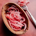 Thinly sliced miso pickled onions in a wooden bowl, with a fork and more onions in the background on a pink surface.