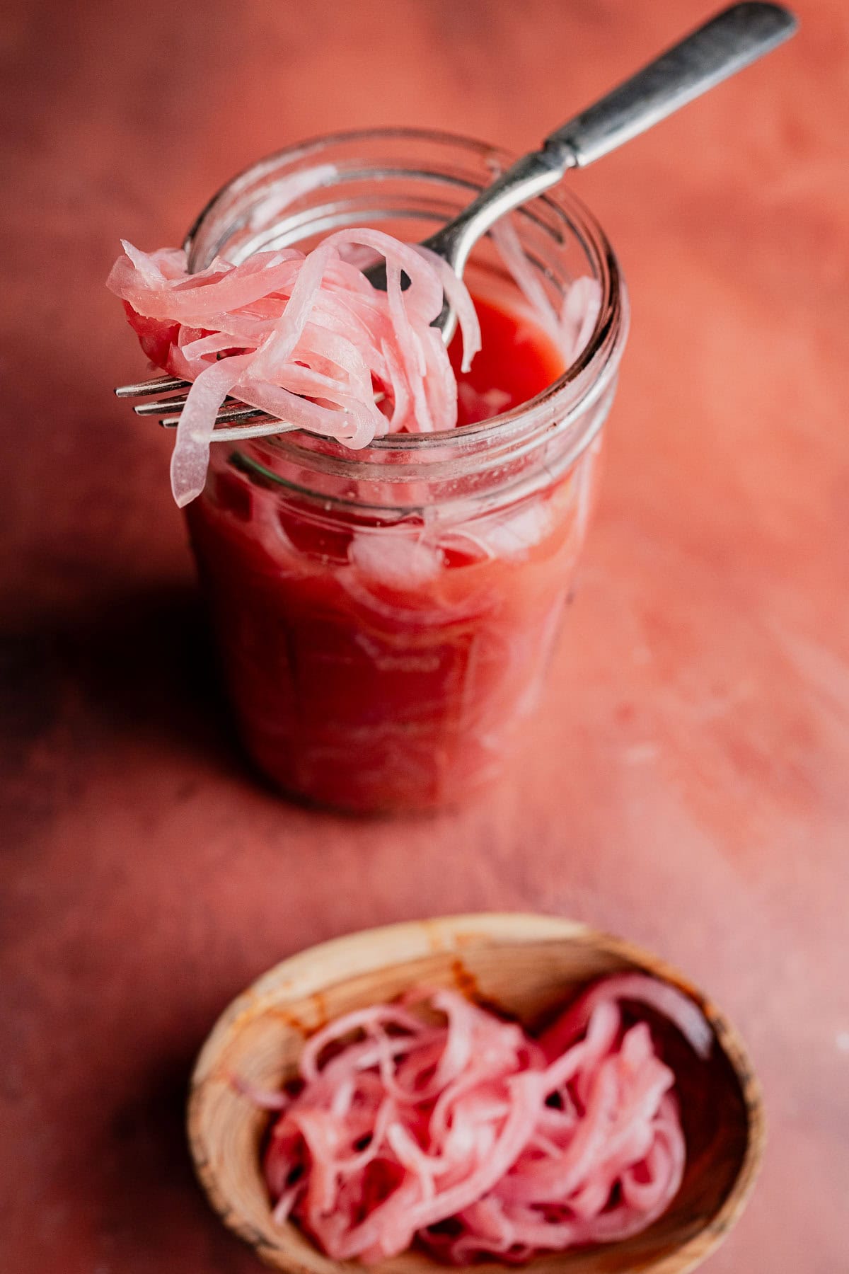 A jar of miso pickled onions with a fork lifting some onions above the jar, and a small dish of onions beside it on a reddish surface.