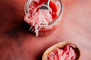 A fork lifts miso pickled onions out of a glass jar, with more tangy pickled onions in a small dish on a reddish surface.