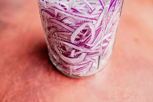 Close-up of thinly sliced miso pickled onions in a glass jar on a reddish surface.
