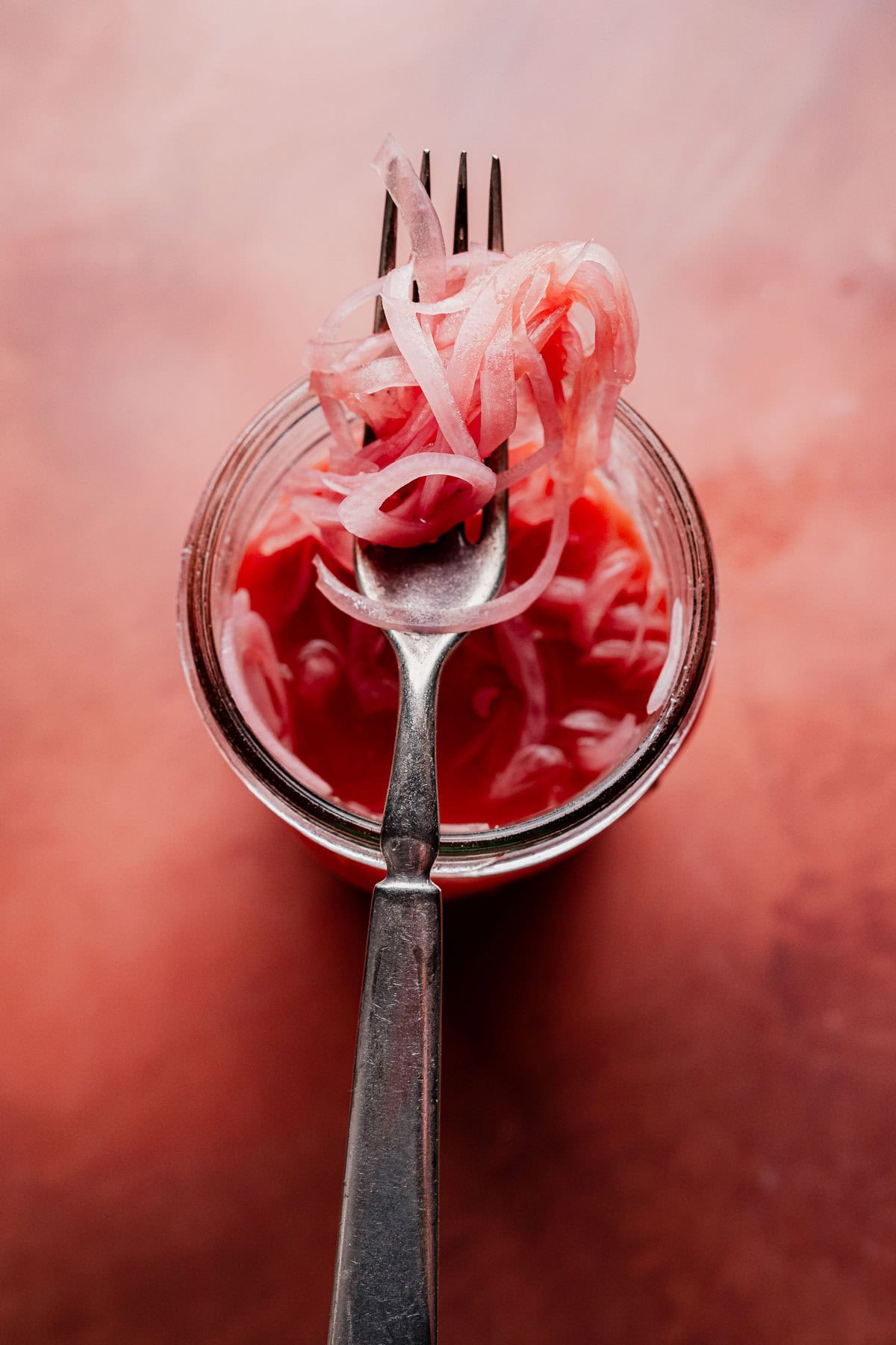 A fork holds up miso pickled onions above a glass jar filled with more onions and tangy pink brine, set against a reddish background.
