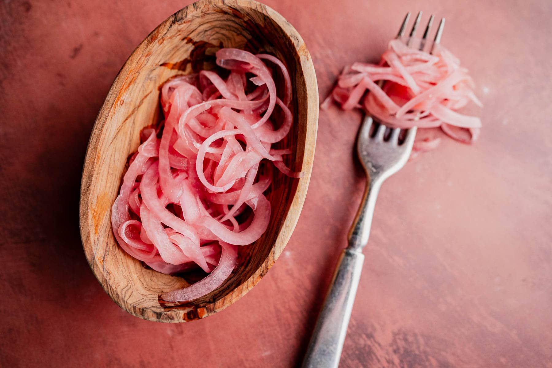 A wooden bowl contains sliced miso pickled onions, with a fork holding more pickled onions on a reddish surface.
