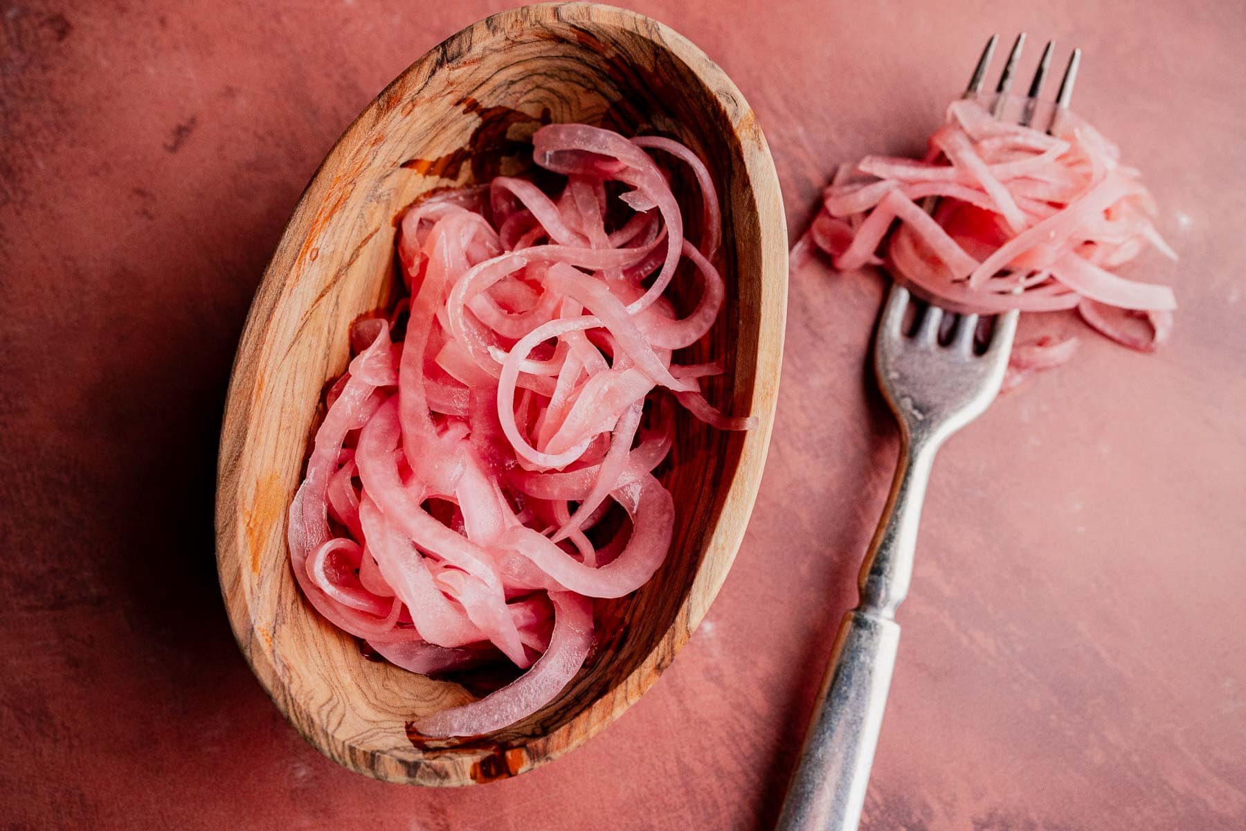 A wooden bowl filled with thinly sliced miso pickled onions, with some onions on a fork beside the bowl, on a pink surface.
