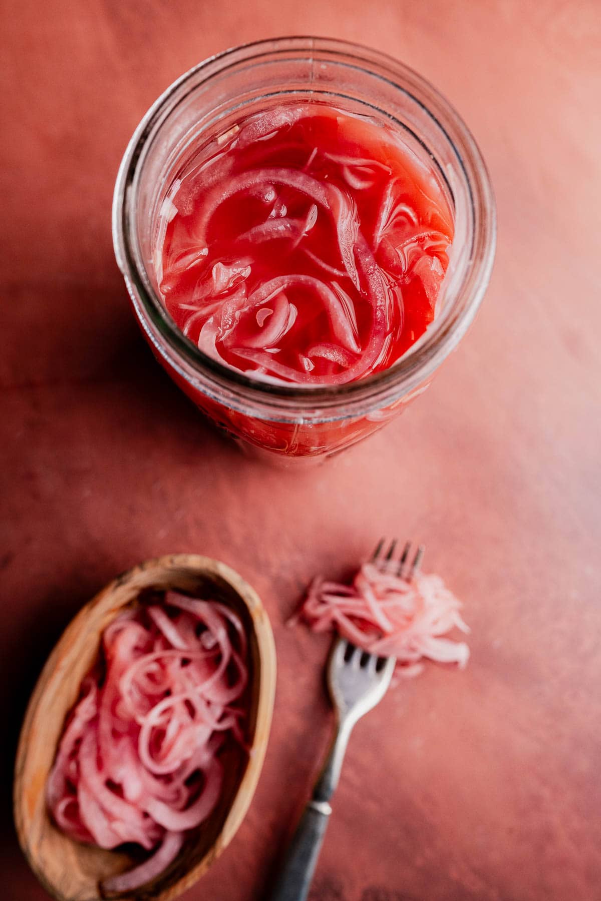A glass jar filled with miso pickled onions on a pink surface, with a small wooden bowl of onions and a fork beside it.