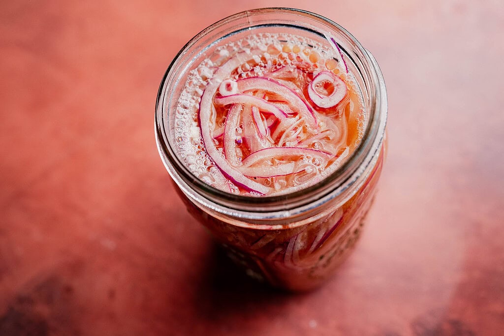 A glass jar filled with miso pickled onions, their vibrant slices soaking in a tangy pickling liquid, rests on a reddish-brown surface.