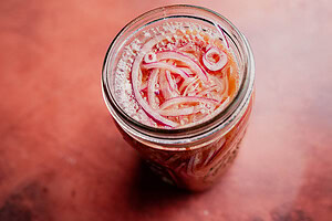 A glass jar filled with miso pickled onions, their vibrant slices soaking in a tangy pickling liquid, rests on a reddish-brown surface.