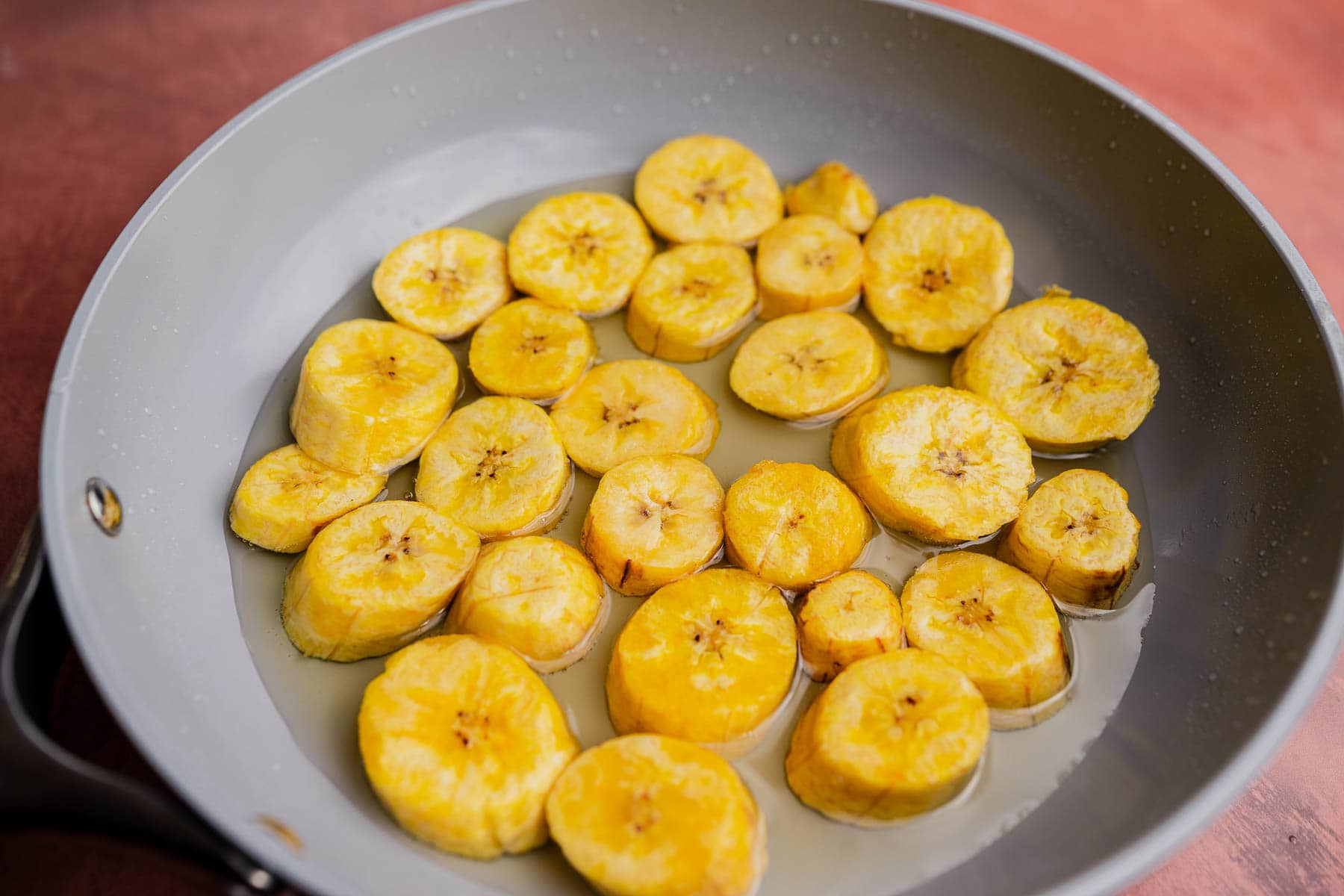 Sliced plantains frying in oil in a gray skillet, perfect for making crispy patacones.