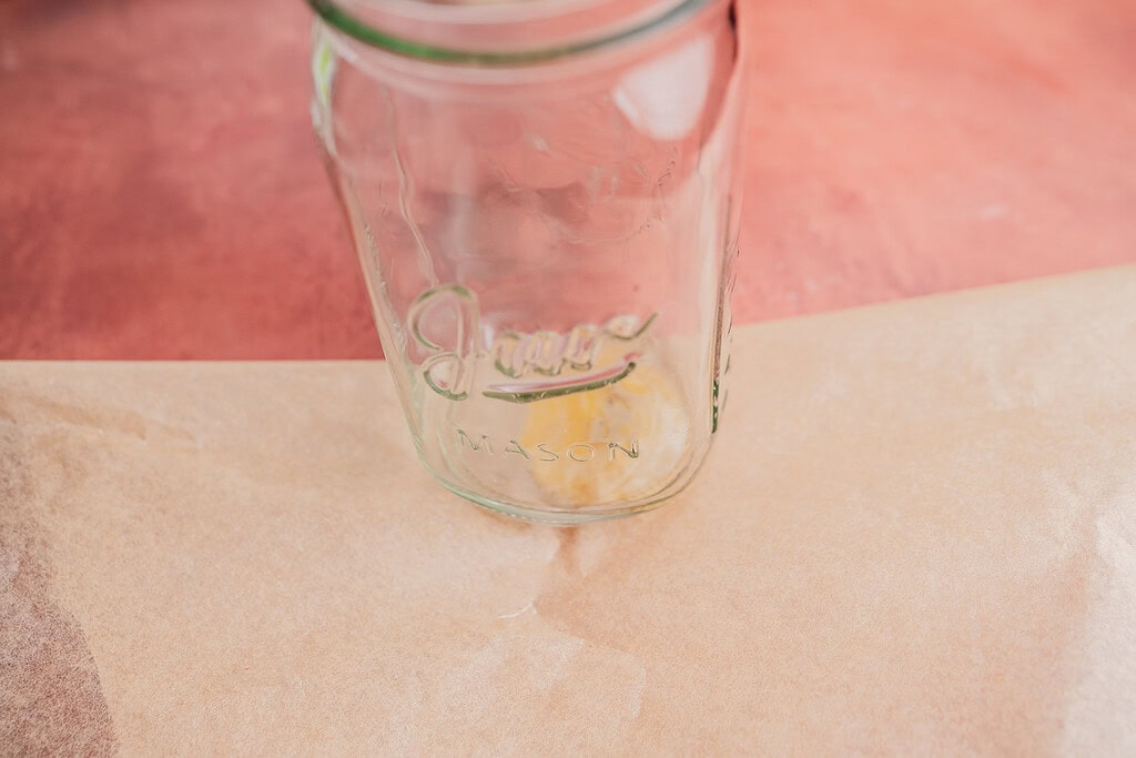 An empty glass mason jar sits on a sheet of brown parchment paper against a pinkish-red background, ready to be filled with freshly made patacones.