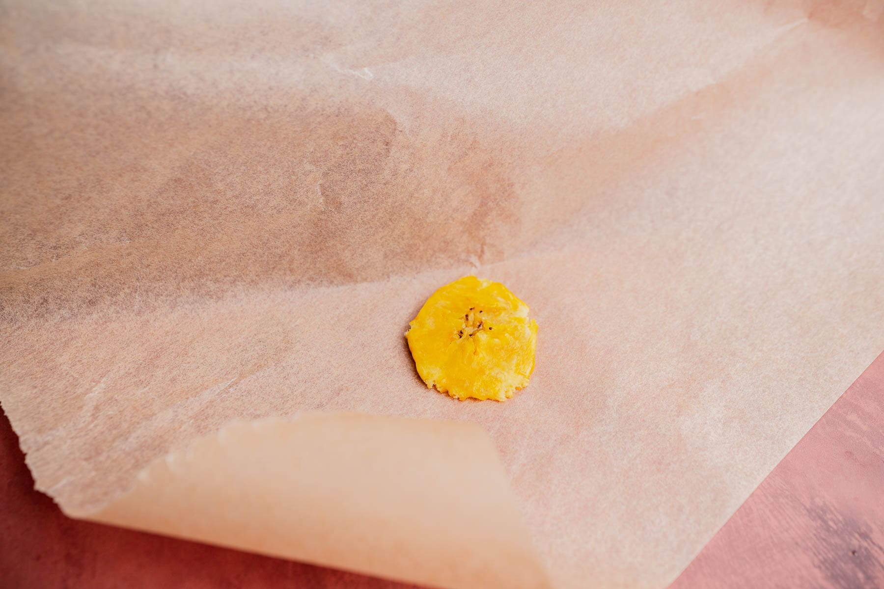A single round yellow patacón dough piece sits on brown parchment paper, with part of the paper folded over it on a pink surface.