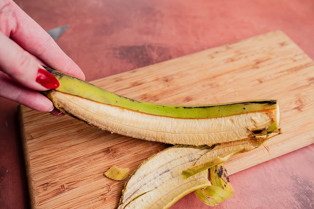 A hand holding a partially peeled plantain over a wooden cutting board, ready to be sliced for patacones, with plantain skin and knife visible in the background.