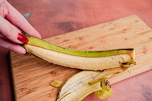 A hand holding a partially peeled plantain over a wooden cutting board, ready to be sliced for patacones, with plantain skin and knife visible in the background.