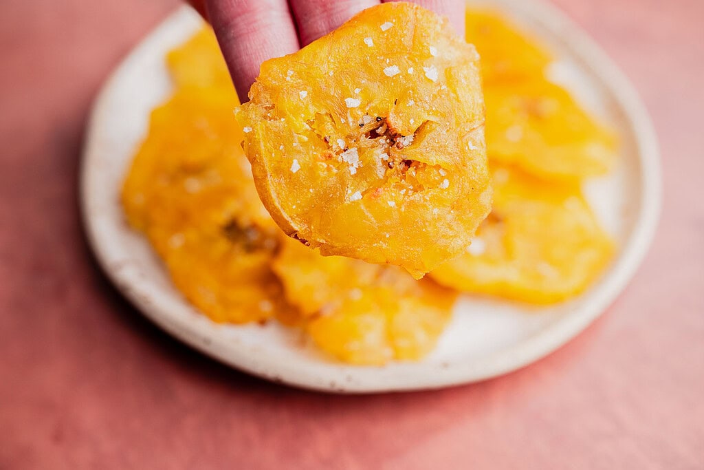 A hand holds a salted, crispy yellow patacones chip above a plate filled with more chips on a pinkish surface.
