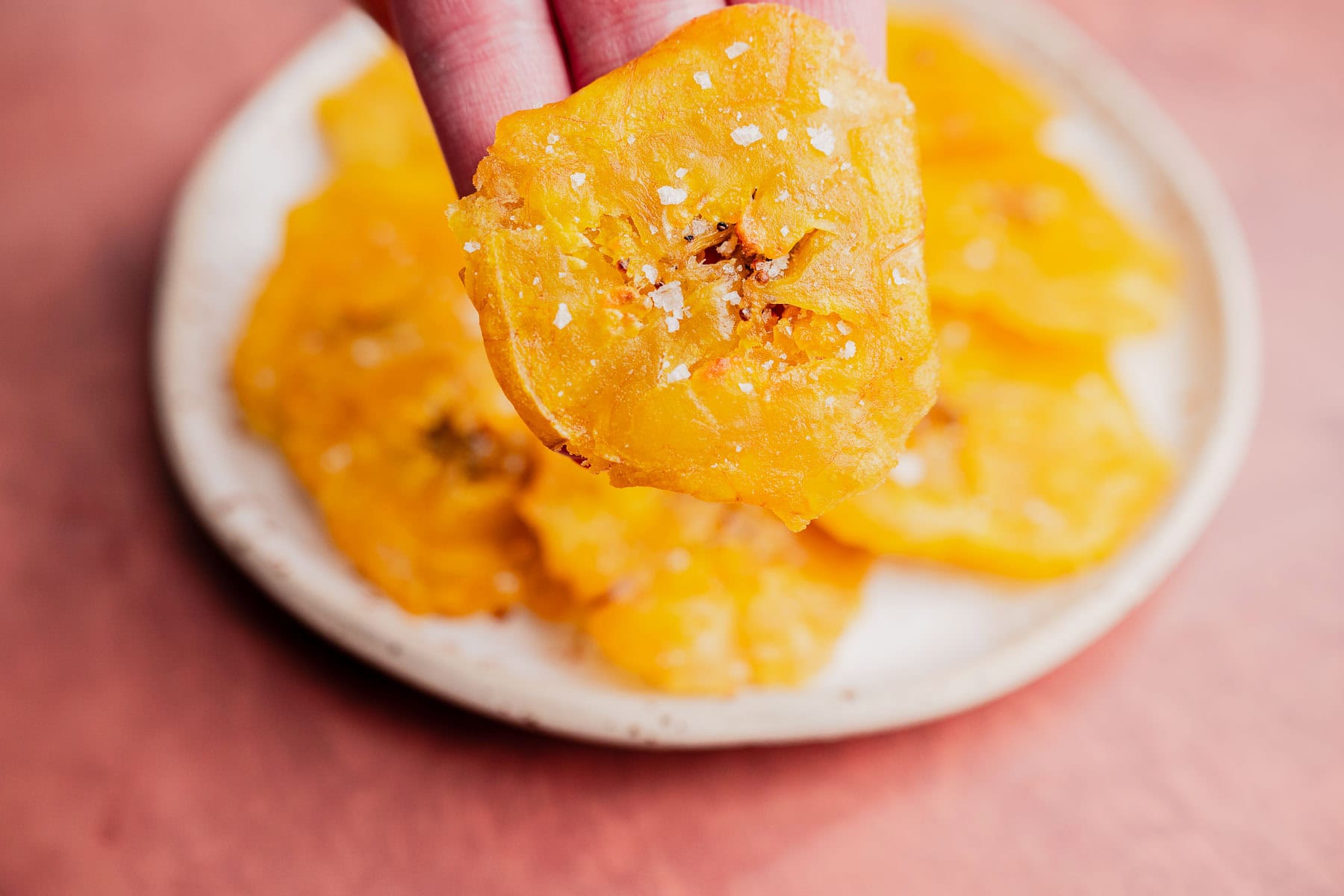 A hand holds a salted, crispy yellow patacones chip above a plate filled with more chips on a pinkish surface.
